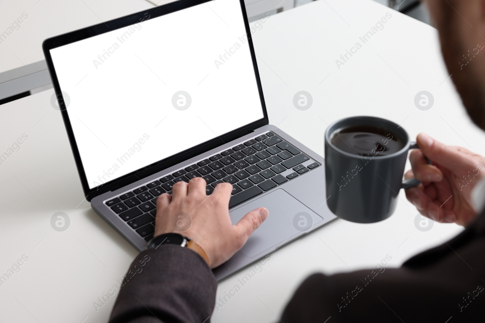 Photo of Man with cup of coffee working on laptop at white desk in office, closeup