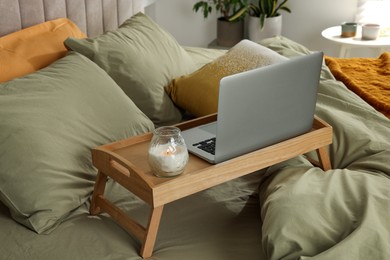 Photo of Wooden tray with modern laptop and burning candle on bed indoors