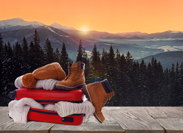 Image of Suitcase with warm clothes on wooden surface against beautiful mountain landscape. Space for text