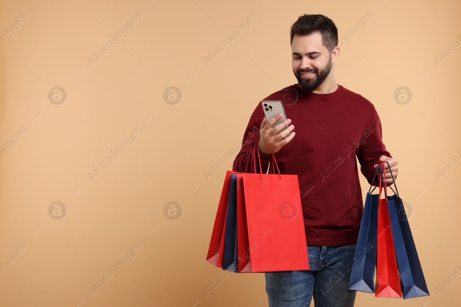 Photo of Smiling man with many paper shopping bags looking at smartphone on beige background. Space for text