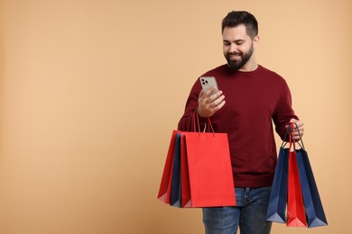 Photo of Smiling man with many paper shopping bags looking at smartphone on beige background. Space for text