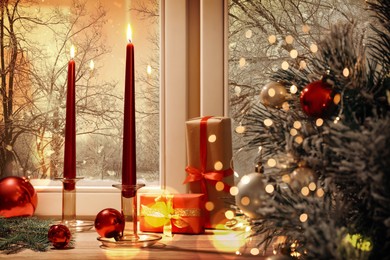 Image of Burning candles, gift boxes and festive decor on window sill near Christmas tree indoors