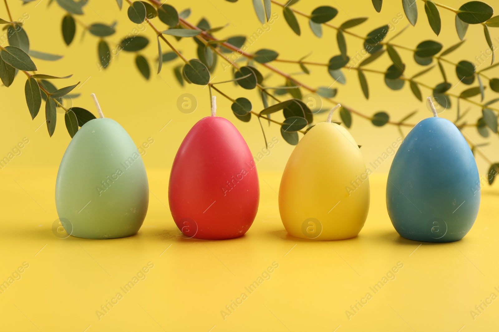 Photo of Colorful egg shaped candles and leaves on yellow background. Easter decor