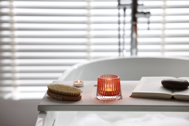 Photo of White wooden tray with burning candles, brush and book on bathtub in bathroom
