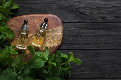 Photo of Bottles of mint essential oil and green leaves on black wooden table, flat lay. Space for text