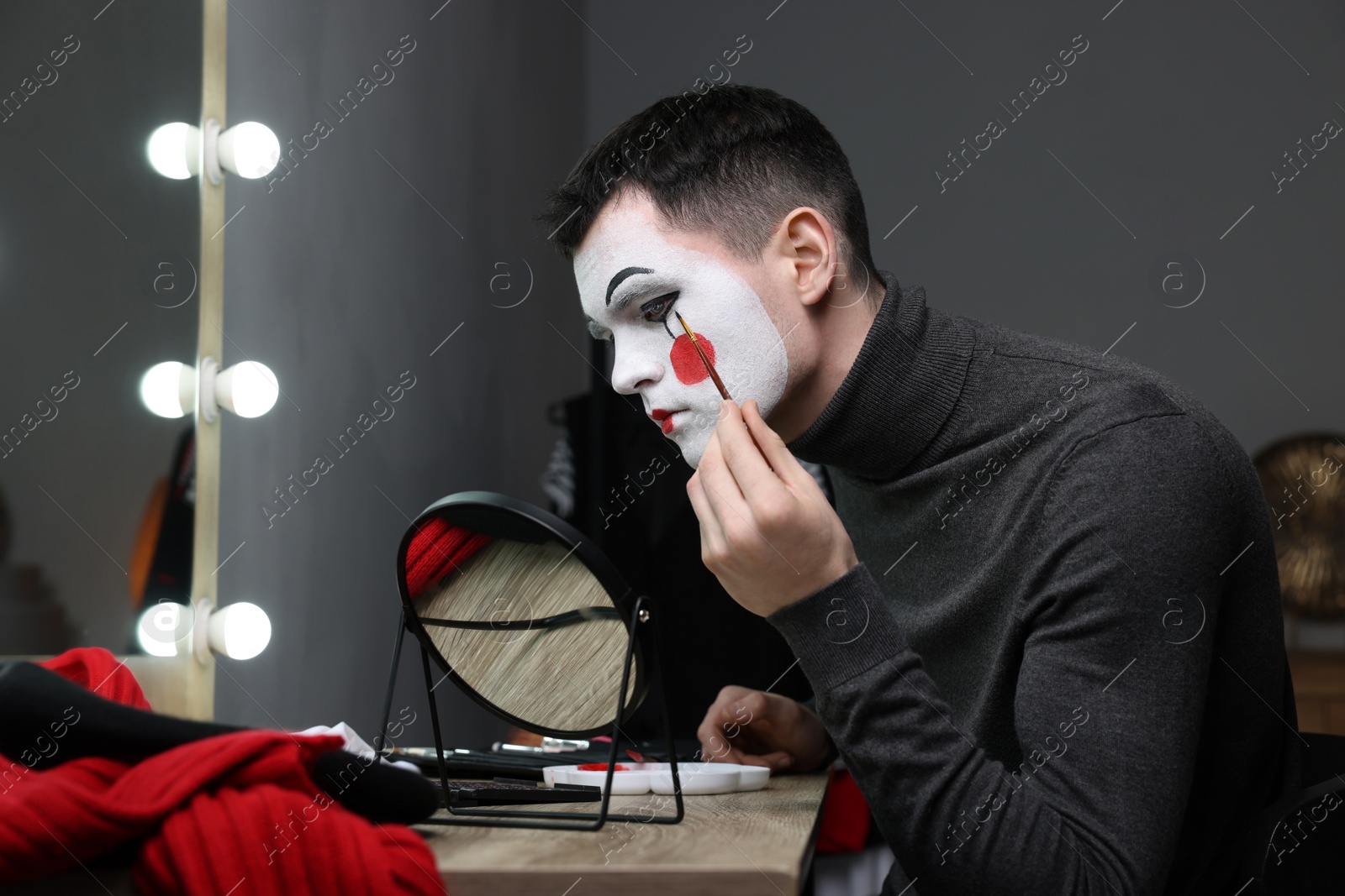 Photo of Young man applying mime makeup near mirror in dressing room