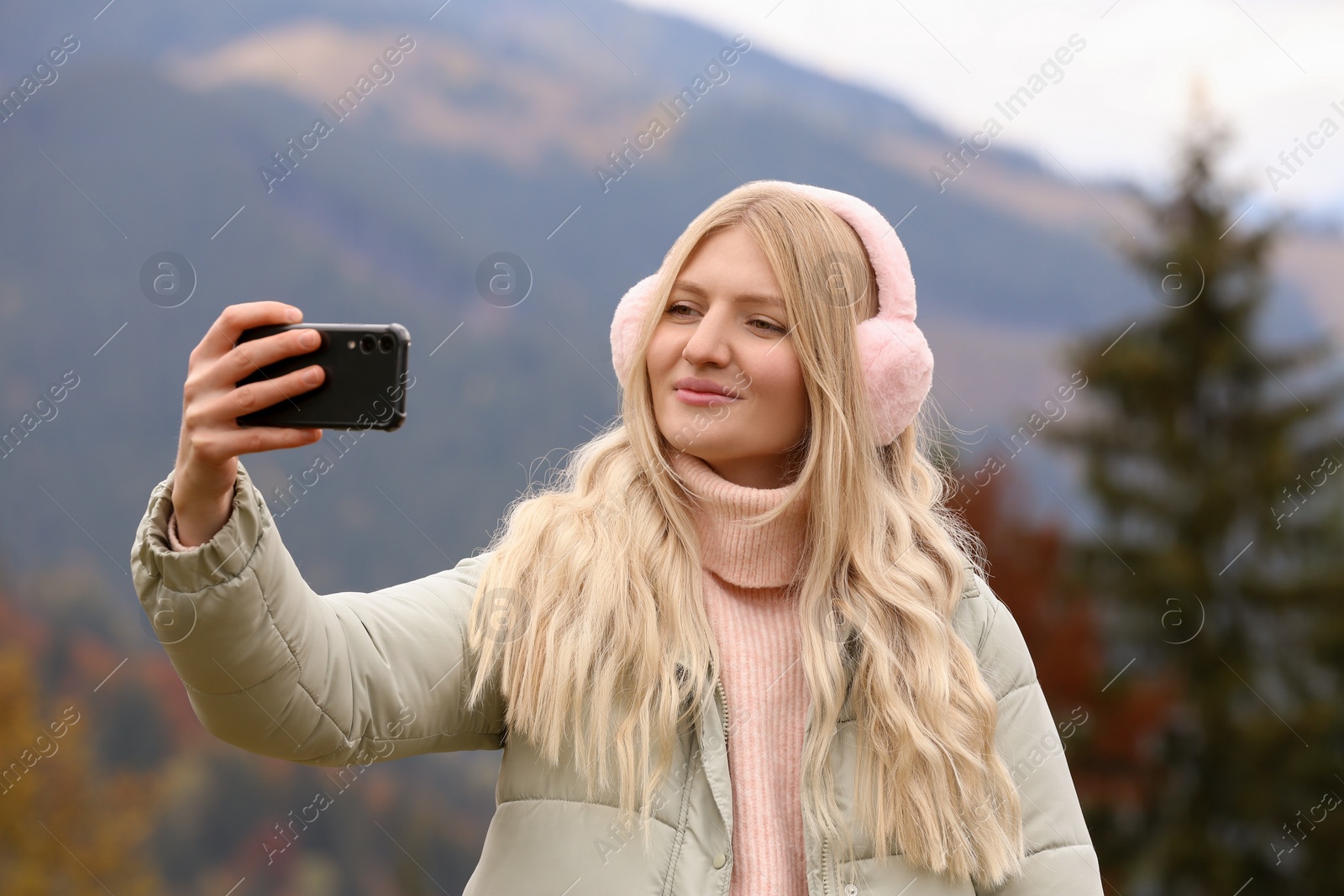 Photo of Young woman in warm earmuffs taking selfie on autumn day