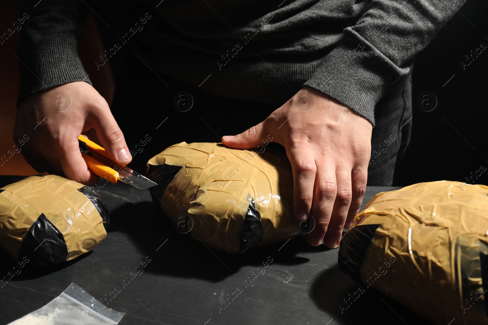 Photo of Smuggling and drug trafficking. Man opening package of narcotics with box cutter at dark table, closeup