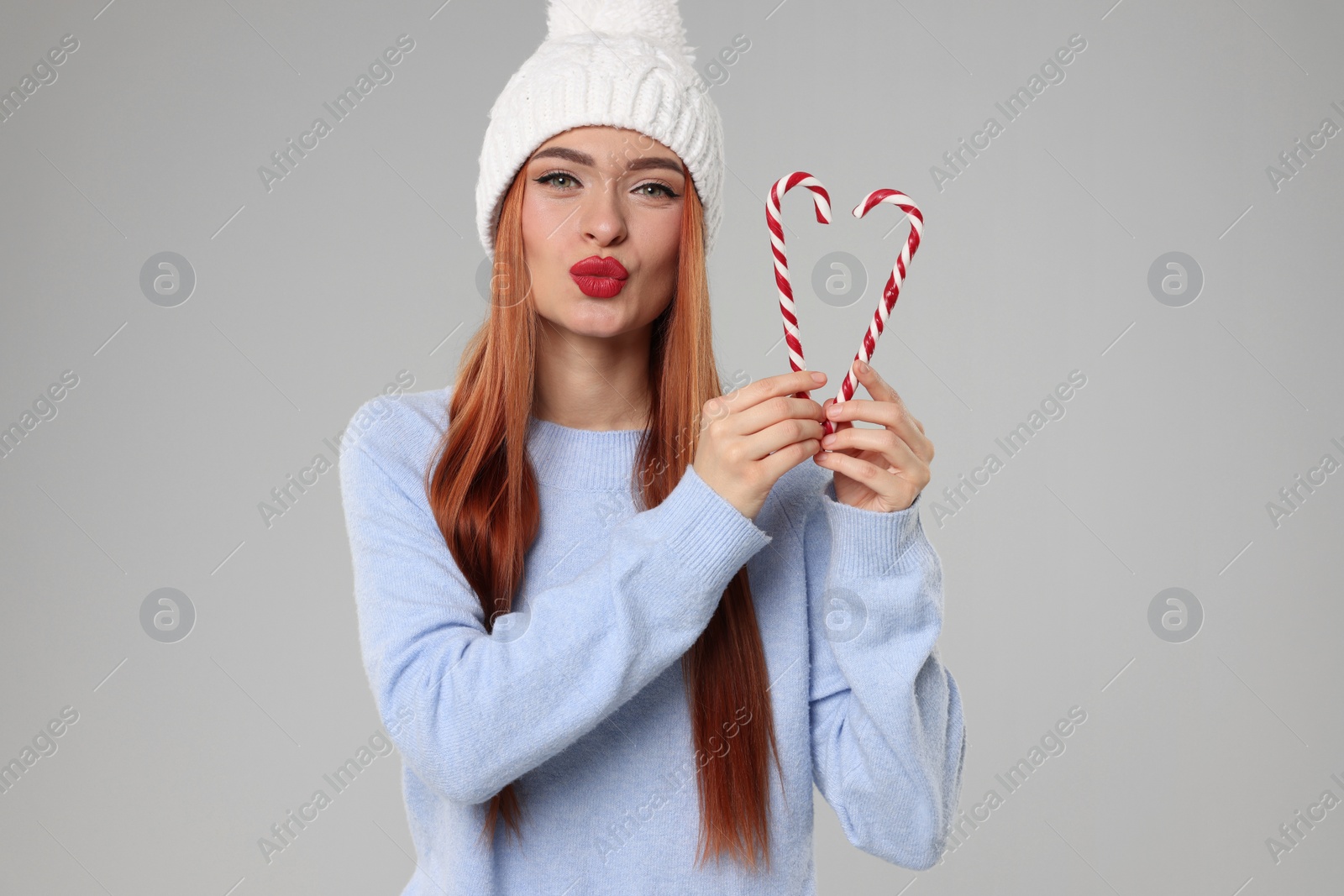 Photo of Young woman in hat and sweater with candy canes on light grey background. Christmas celebration