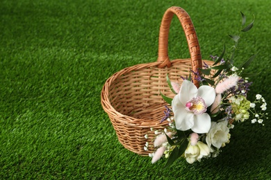 Wicker basket decorated with beautiful flowers on green grass, space for text. Easter item