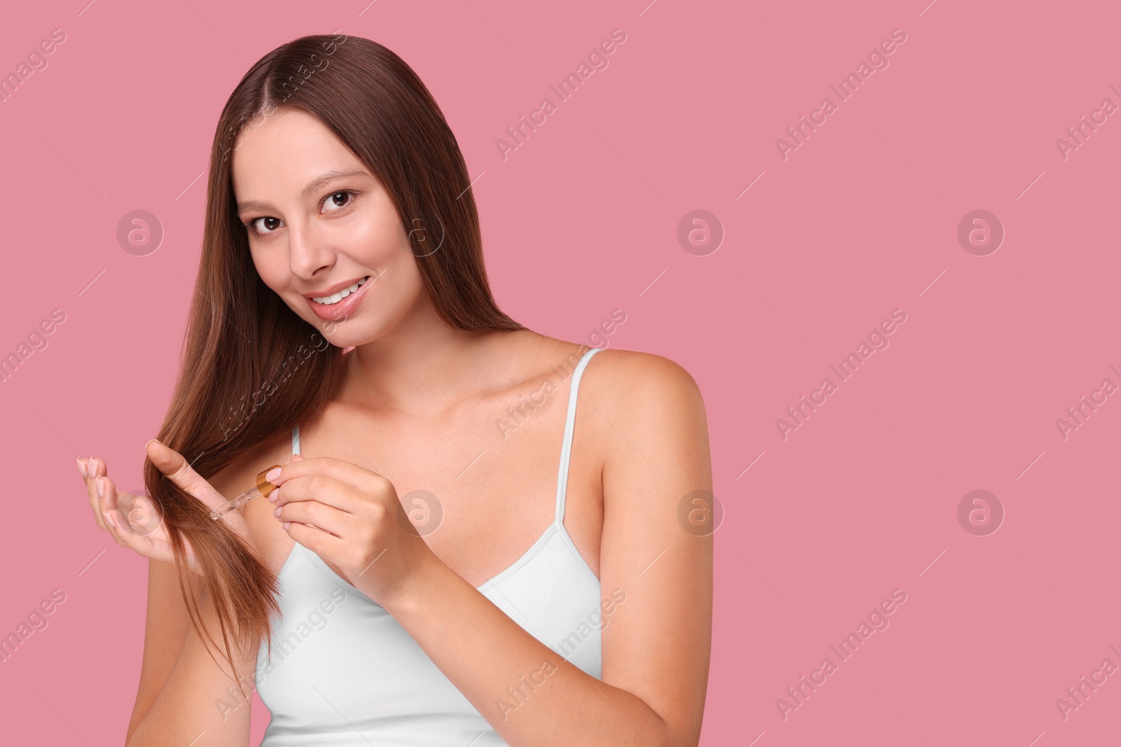 Photo of Beautiful woman applying serum onto hair on pink background, space for text