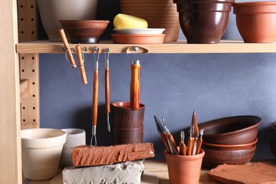 Photo of Set of different crafting tools and clay dishes on wooden rack in workshop, closeup