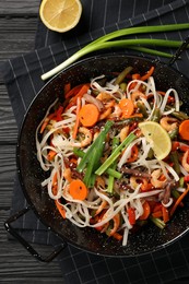 Photo of Shrimp stir fry with noodles and vegetables in wok on black wooden table, top view