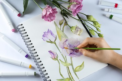 Woman painting eustomas in sketchbook at white table, top view