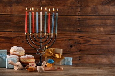 Hanukkah celebration. Menorah with burning candles, dreidels, donuts and gift boxes on wooden table, space for text