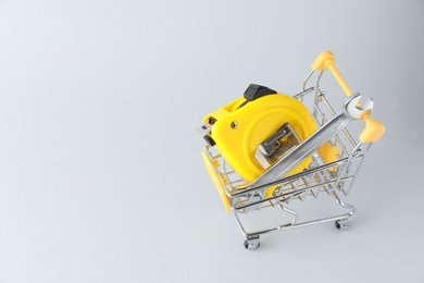 Metal wrench and measuring tape in shopping cart on light grey background, space for text