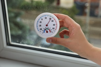 Woman holding round hygrometer with thermometer near window on rainy day, closeup