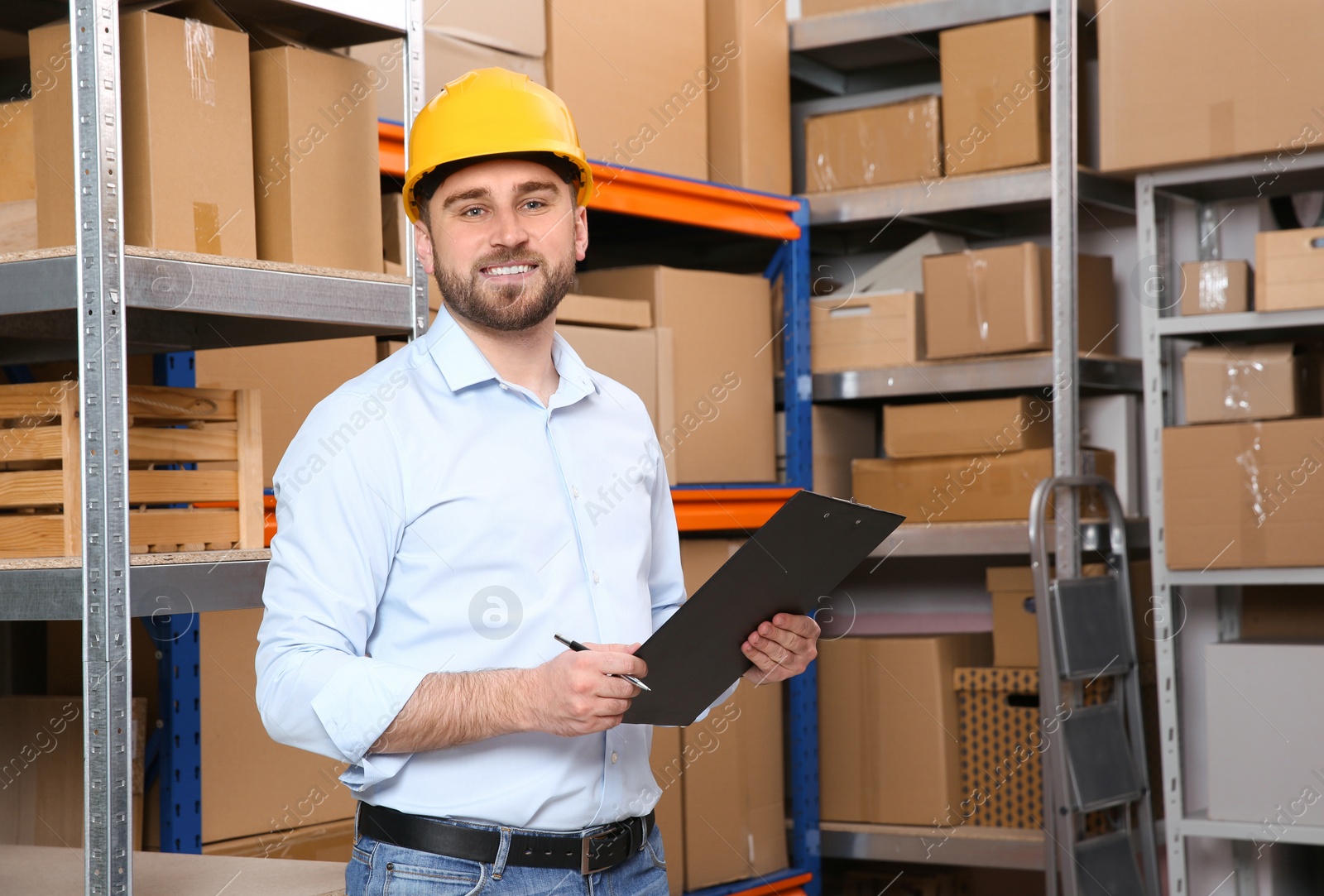 Photo of Young man with clipboard near rack of cardboard boxes at warehouse