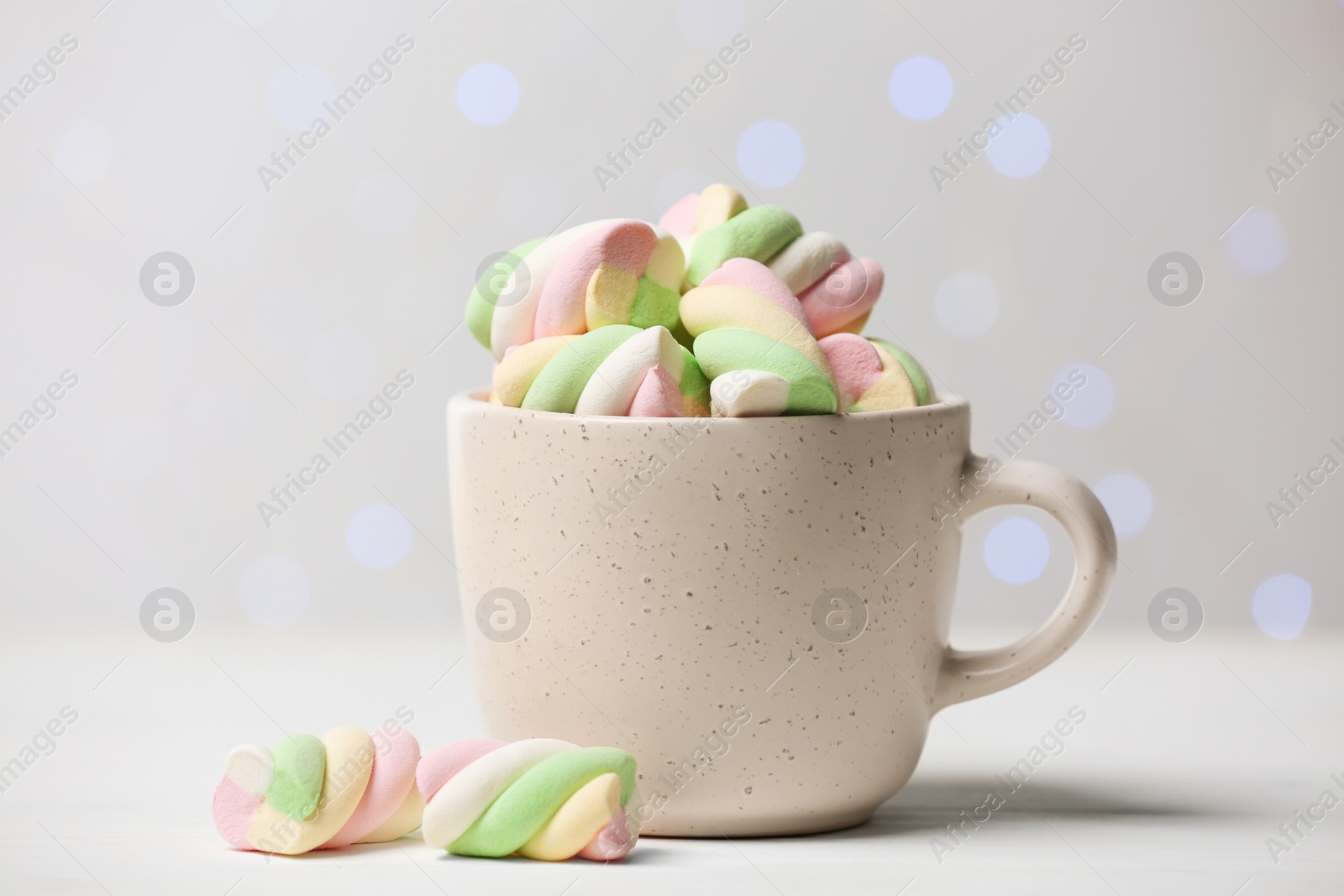 Photo of Ceramic cup with colorful marshmallows on white table against blurred lights