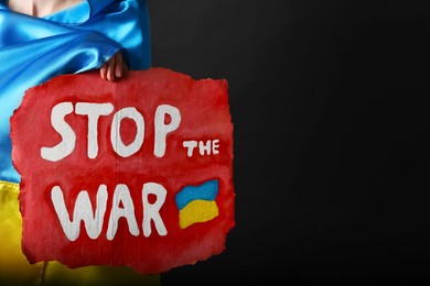 Woman wrapped in Ukrainian flag holding poster with words Stop the War on black background, closeup. Space for text