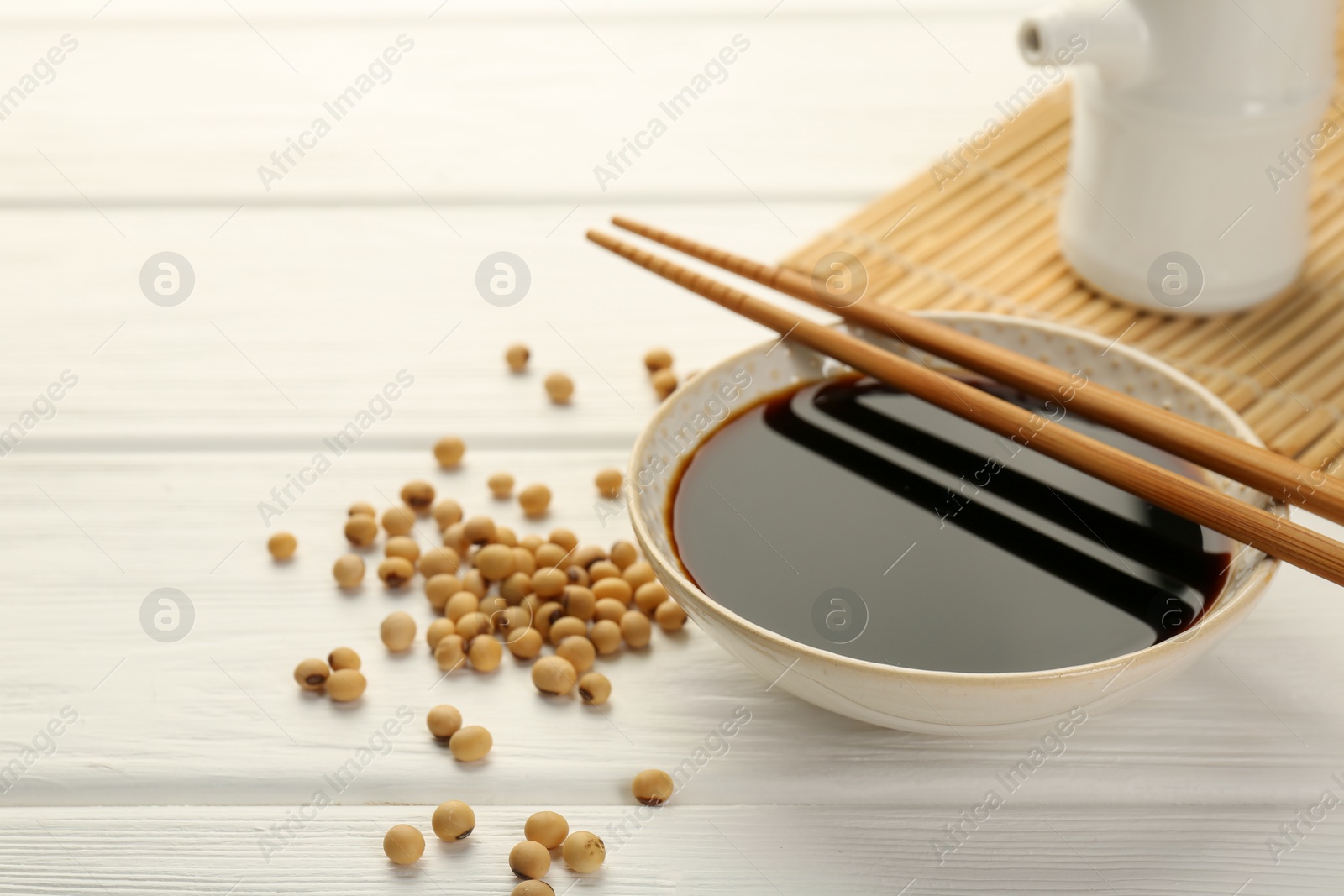 Photo of Tasty soy sauce in bowl, chopsticks and soybeans on white wooden table, closeup