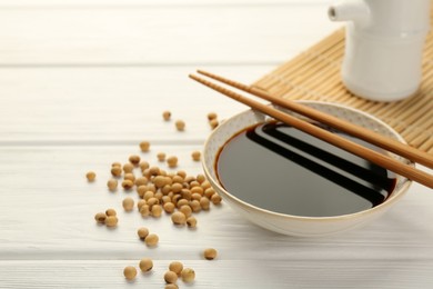 Tasty soy sauce in bowl, chopsticks and soybeans on white wooden table, closeup