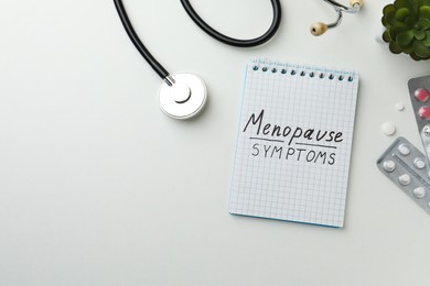 Photo of Notebook with words Menopause Symptoms, pills, stethoscope and plant on white table, flat lay. Space for text