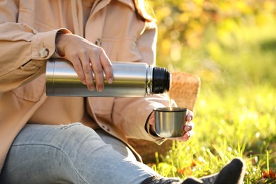 Photo of Woman pouring tea from thermos into cup lid on green grass outdoors, closeup