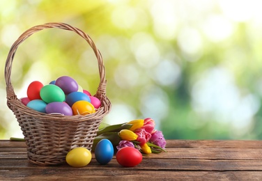 Colorful Easter eggs in wicker basket and tulips on wooden table outdoors, space for text