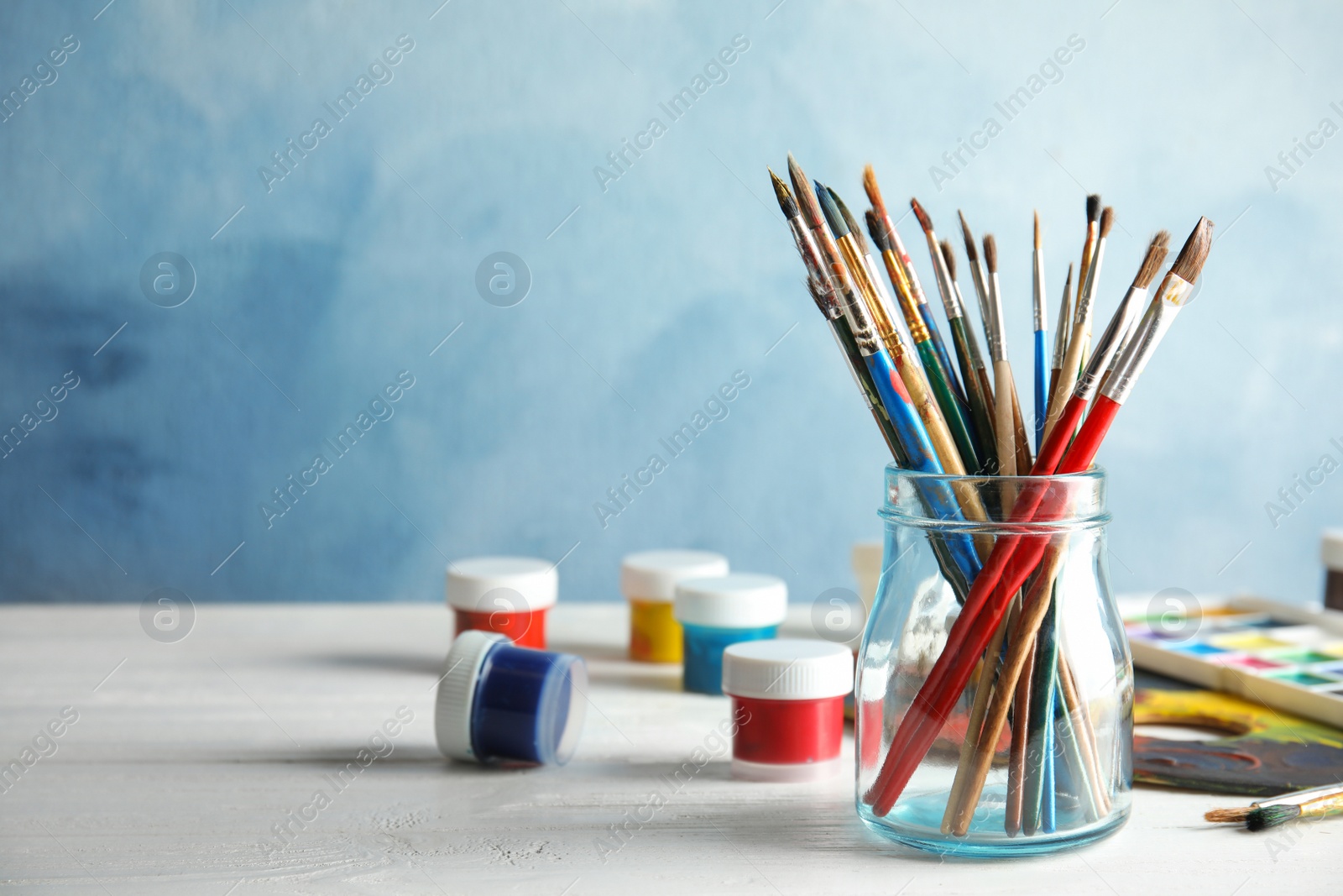 Photo of Glass jar with brushes and paints on table against color background. Space for text