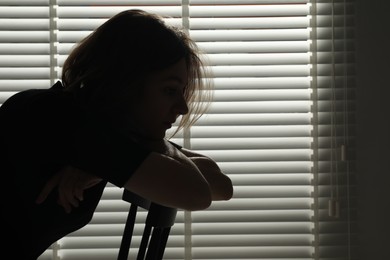 Photo of Silhouette of sad young woman near closed blinds indoors, space for text