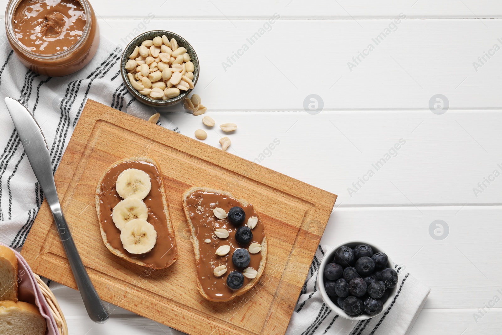 Photo of Toasts with tasty nut butter, banana slices, blueberries and peanuts on white wooden table, flat lay. Space for text