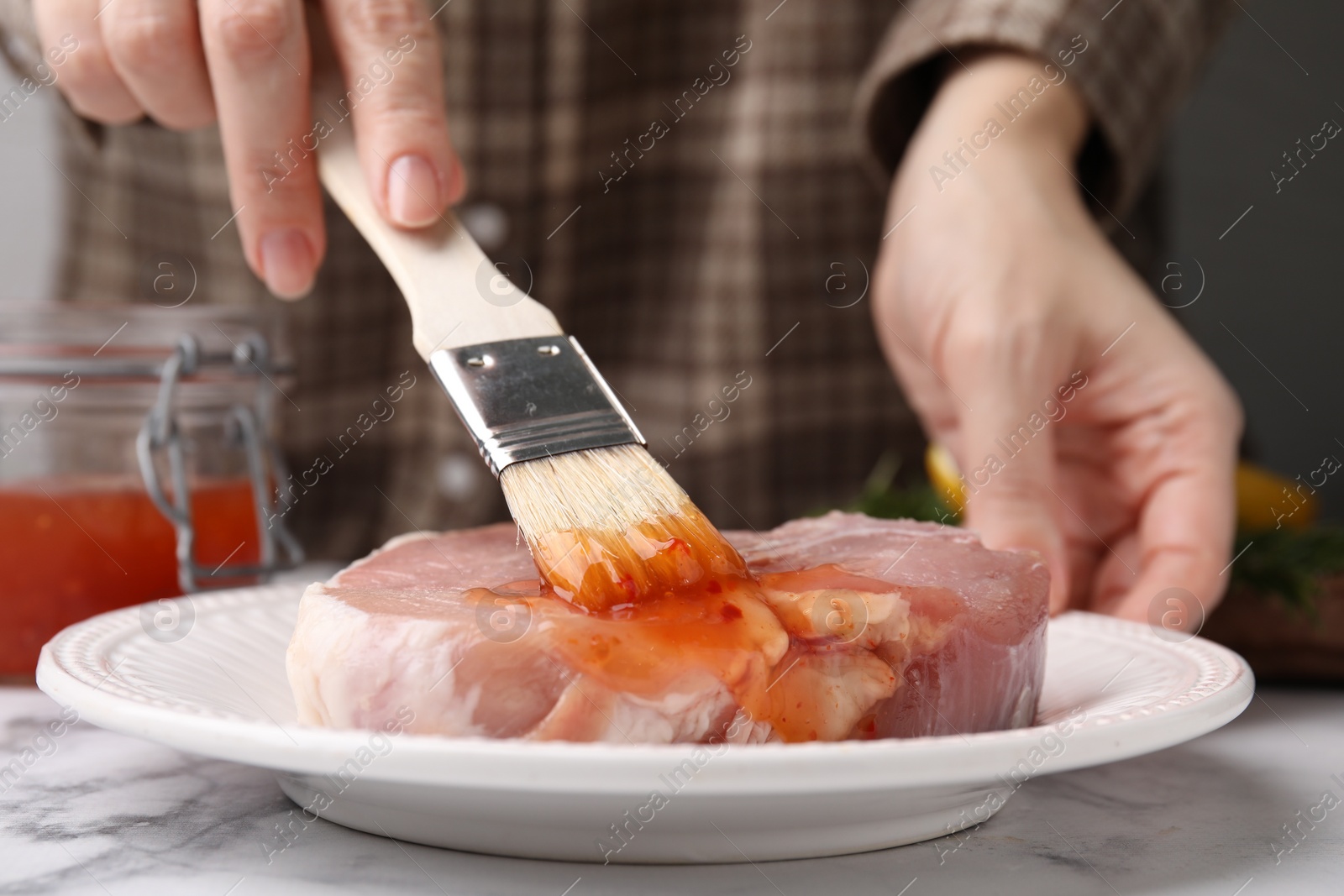 Photo of Woman spreading marinade onto raw meat with basting brush at white marble table, closeup