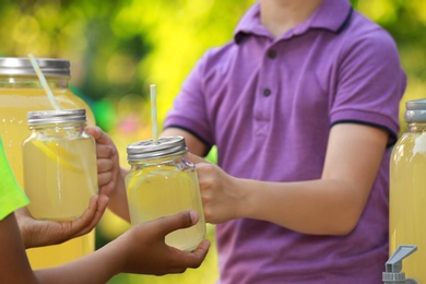 Photo of Little boy selling natural lemonade to African-American kid in park, closeup. Summer refreshing drink