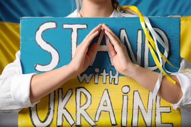 Photo of Woman holding poster with words Stand with Ukraine near national flag, closeup