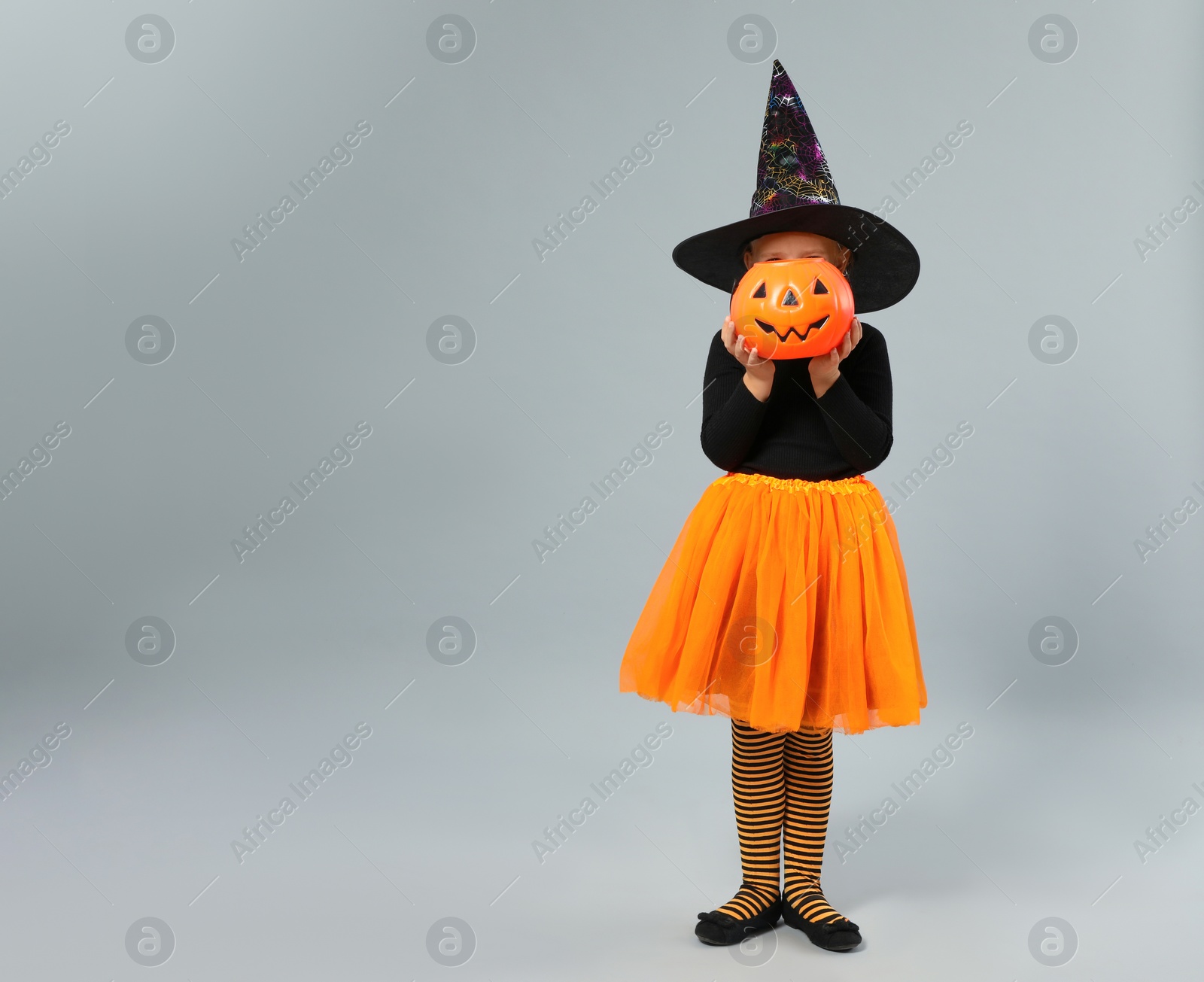 Photo of Cute little girl with pumpkin candy bucket wearing Halloween costume on grey background, space for text