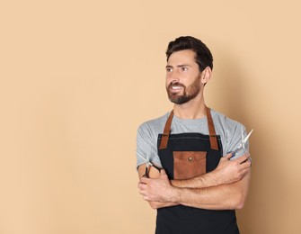 Smiling hairdresser in apron holding scissors on light brown background, space for text