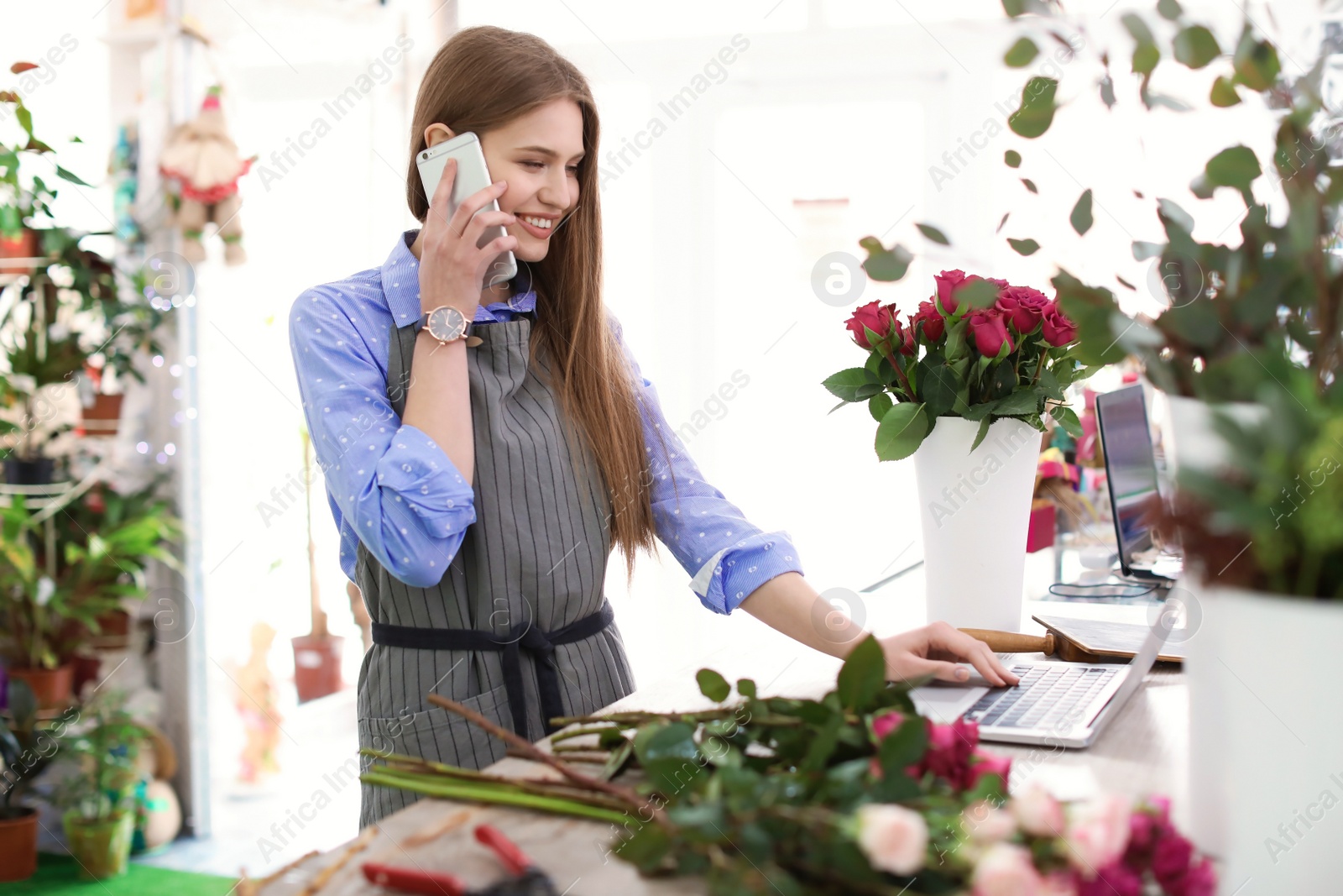 Photo of Young florist working with laptop and talking on mobile phone in flower shop