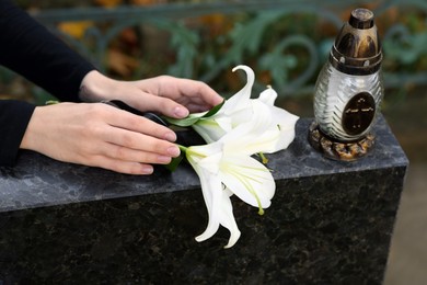 Photo of Woman putting white lilies and grave lantern on tombstone at cemetery, closeup