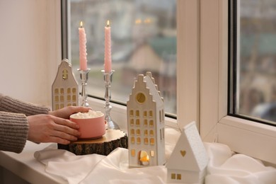 Woman holding cup of delicious drink near windowsill with house shaped lanterns indoors, closeup