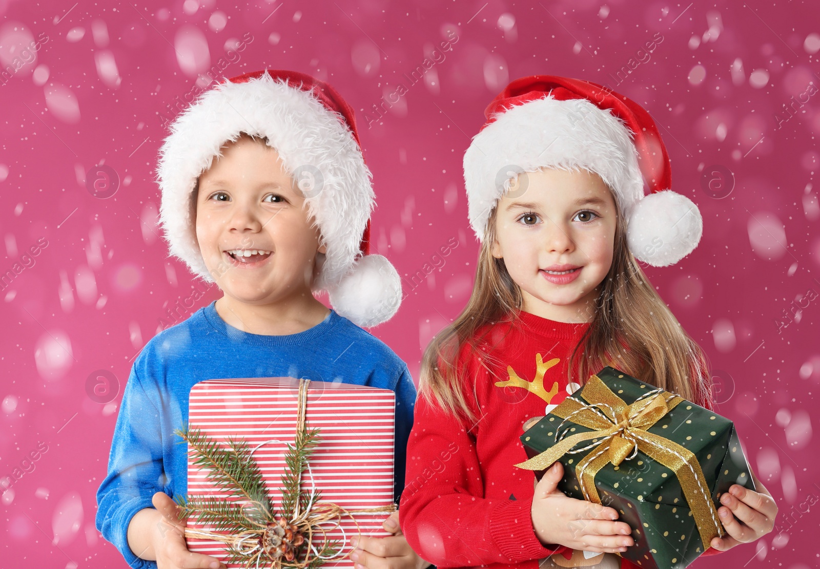 Image of Cute children in Santa hats with Christmas gifts under snowfall on pink background