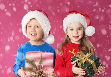 Cute children in Santa hats with Christmas gifts under snowfall on pink background