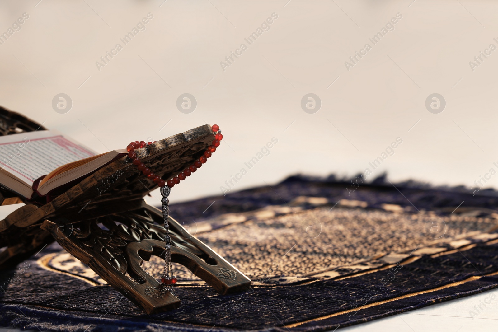 Photo of Rehal with open Quran and Muslim prayer beads on rug indoors. Space for text