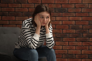 Sad young woman sitting on chair near brick wall, space for text