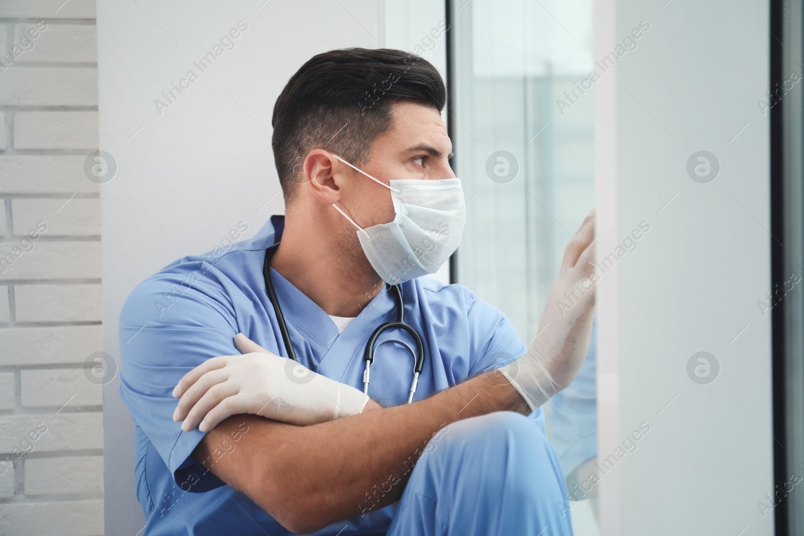 Photo of Sad doctor near window indoors. Stress of health care workers during coronavirus pandemic