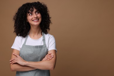 Photo of Happy woman wearing kitchen apron on brown background, space for text. Mockup for design