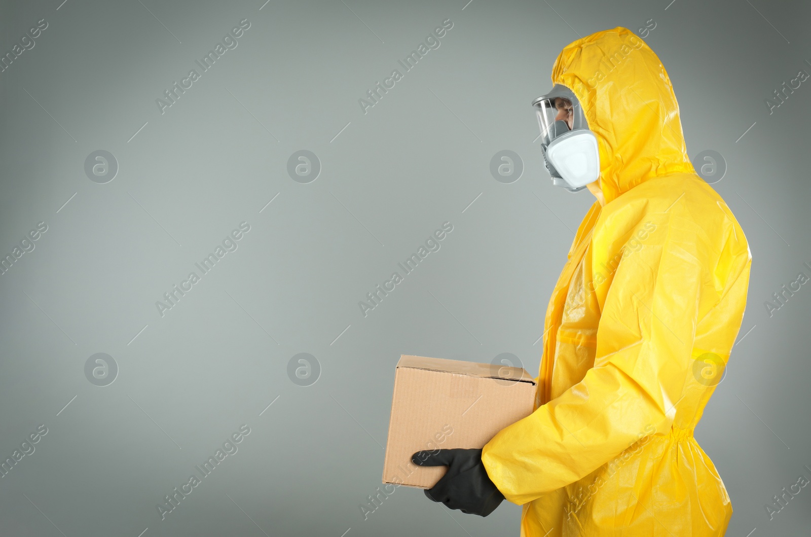 Photo of Man wearing chemical protective suit with cardboard box on light grey background, space for text. Prevention of virus spread