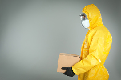 Photo of Man wearing chemical protective suit with cardboard box on light grey background, space for text. Prevention of virus spread