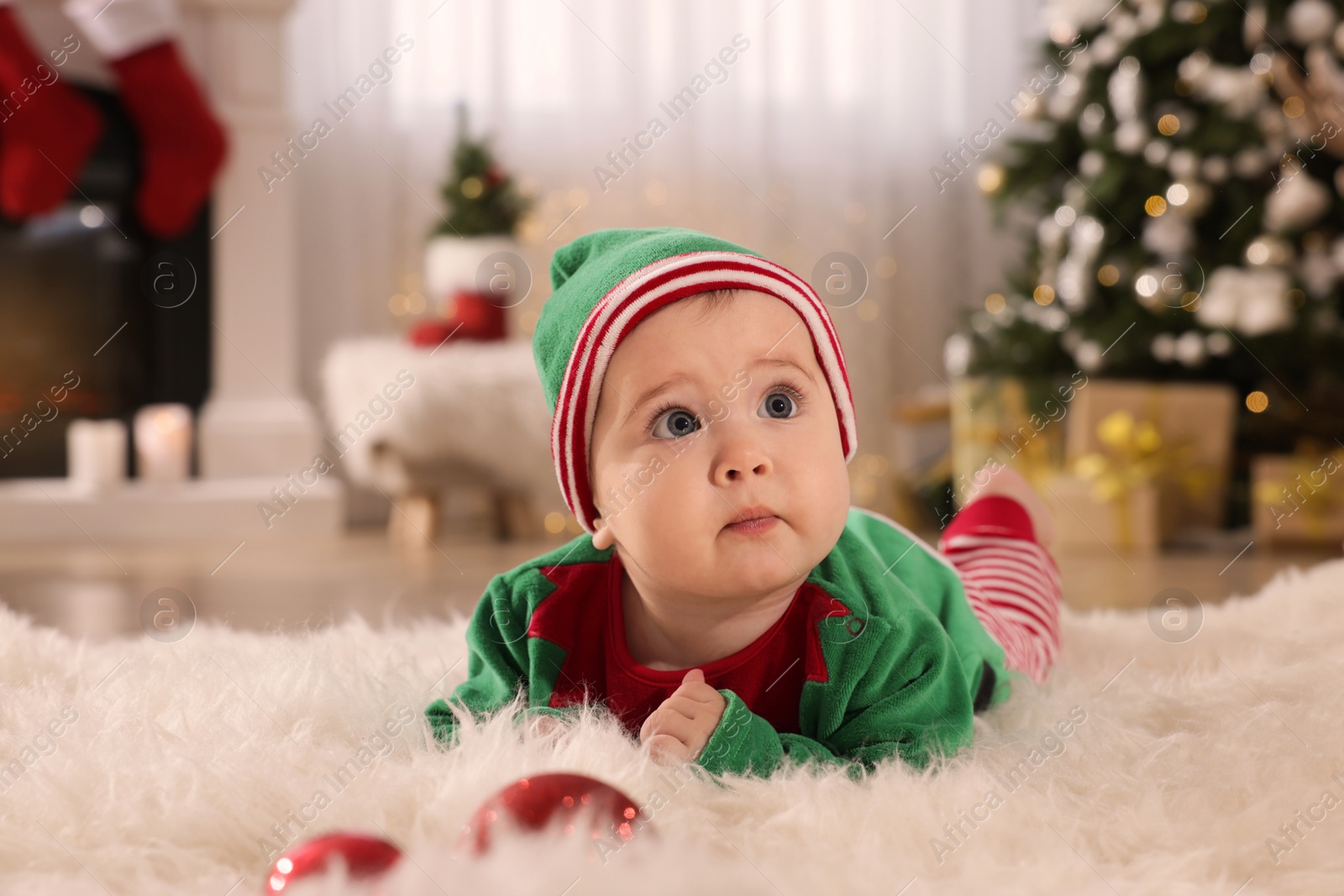 Photo of Baby wearing cute elf costume on floor in room decorated for Christmas