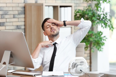 Businessman suffering from heat in front of small fan at workplace